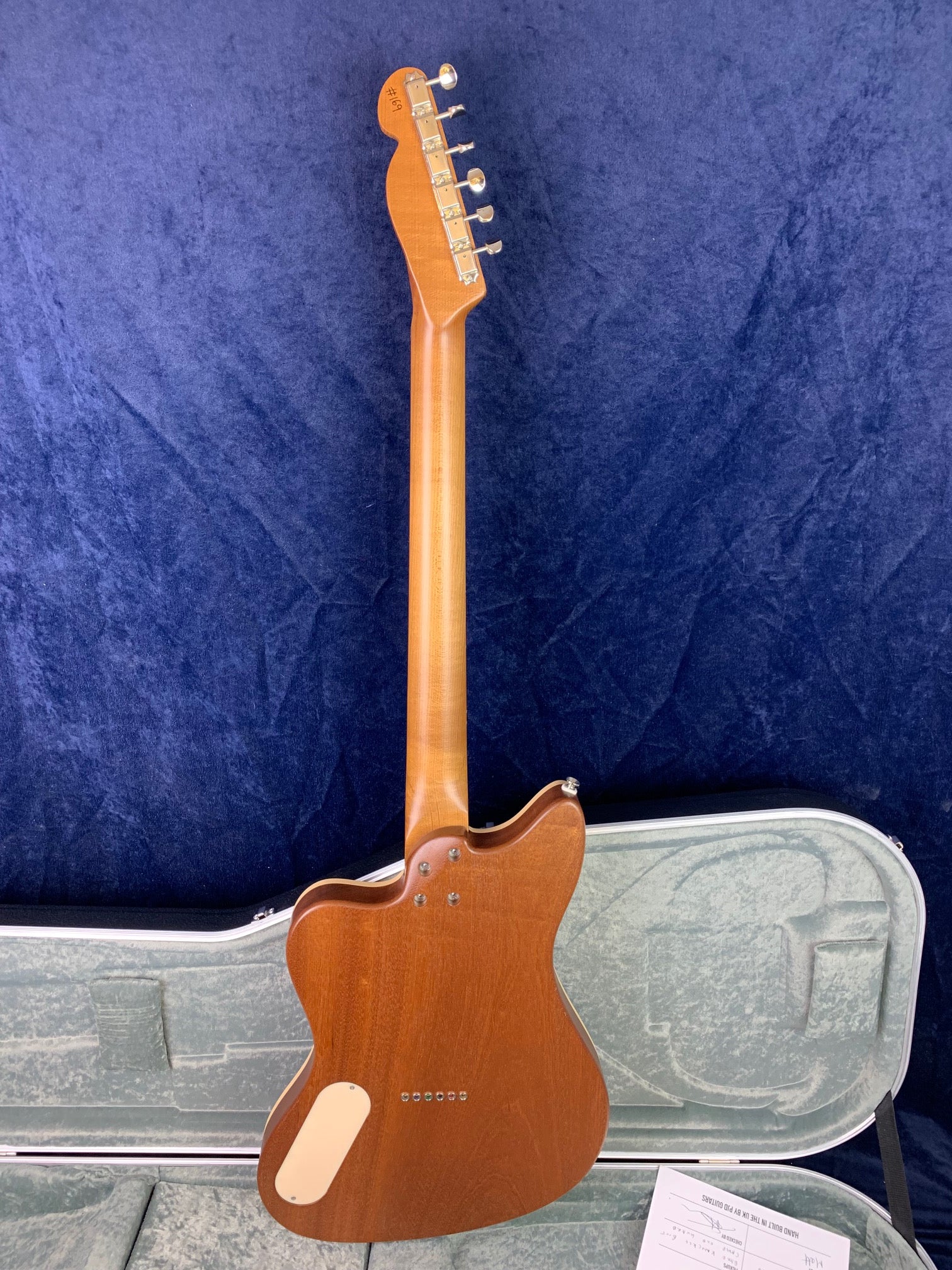 A brown electric guitar seen from the back, showing a single cutaway body, metal neck screws, and light-colored control cover, resting in an open case.