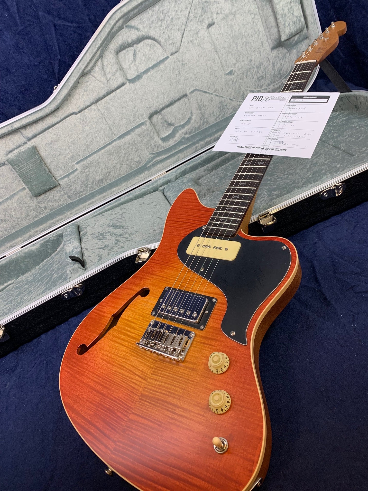 Sunburst electric guitar with a semi-hollow body, two pickups, two control knobs, in an open gray hard case with a spec sheet on the neck.