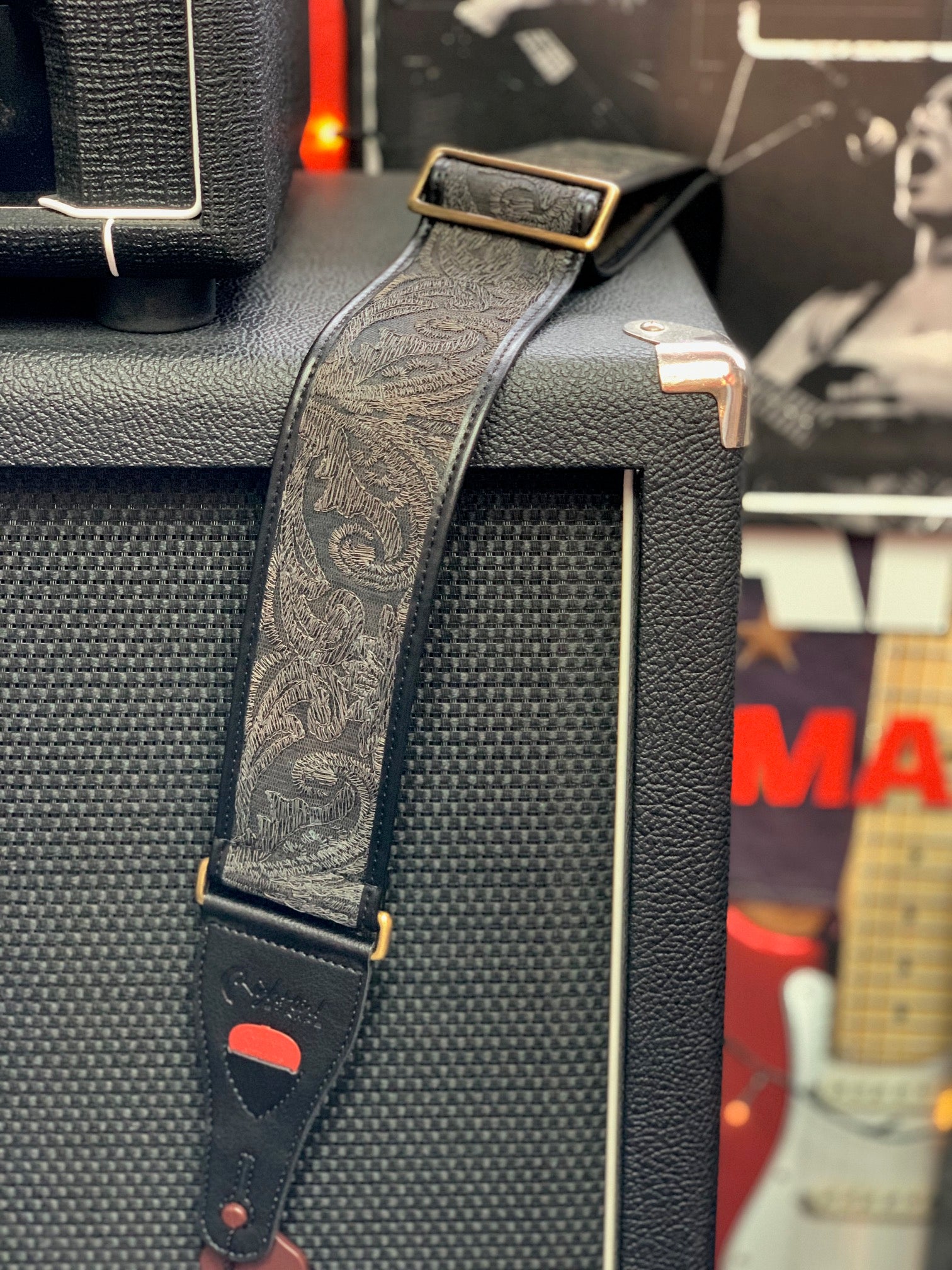 Black guitar strap with paisley pattern rests on textured black amplifier, showing a brass buckle and leather end with a guitar pick holder.