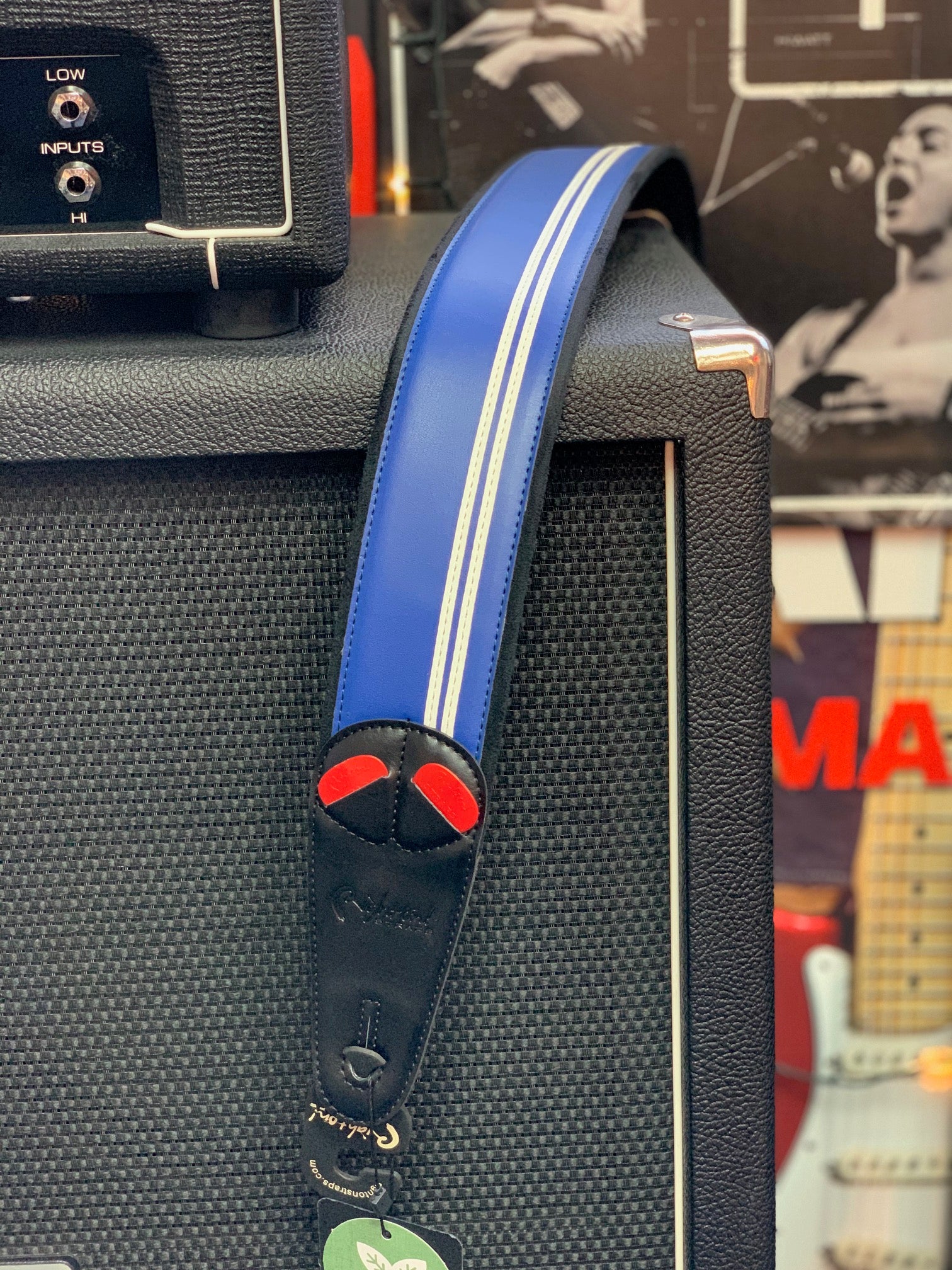 Blue guitar strap with white stripes, black leather ends, and red accents draped over a black amplifier with a textured surface.