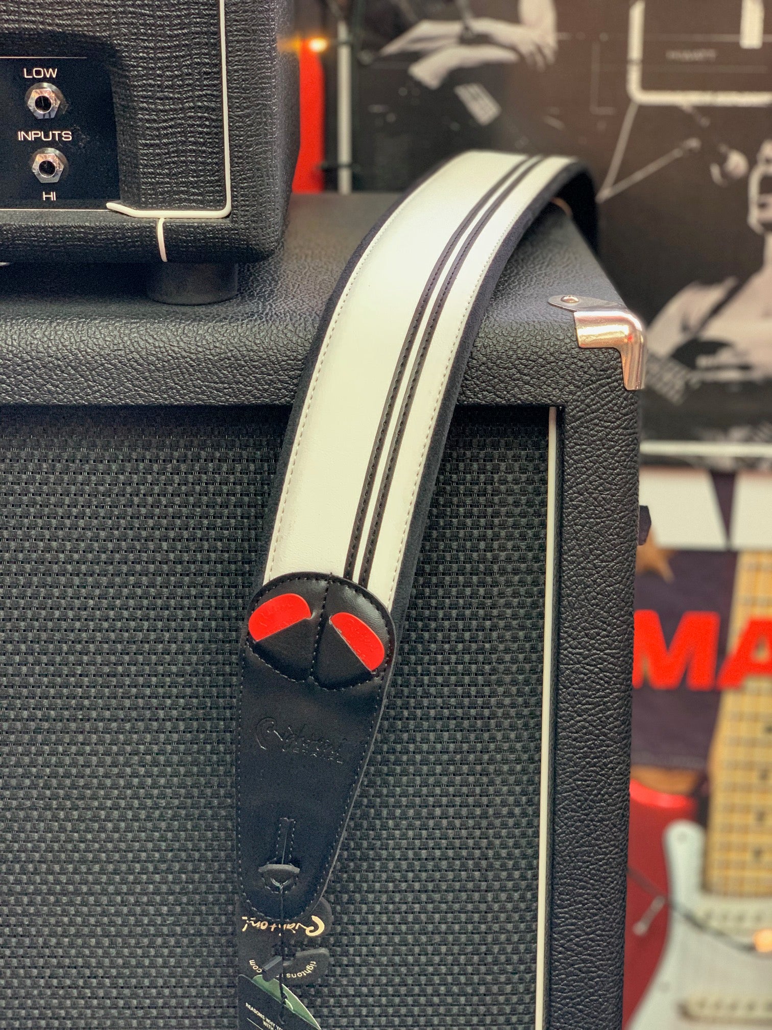 White and black leather guitar strap with red accents draped over a black amplifier cabinet, nearby guitar and amp details visible in the background.