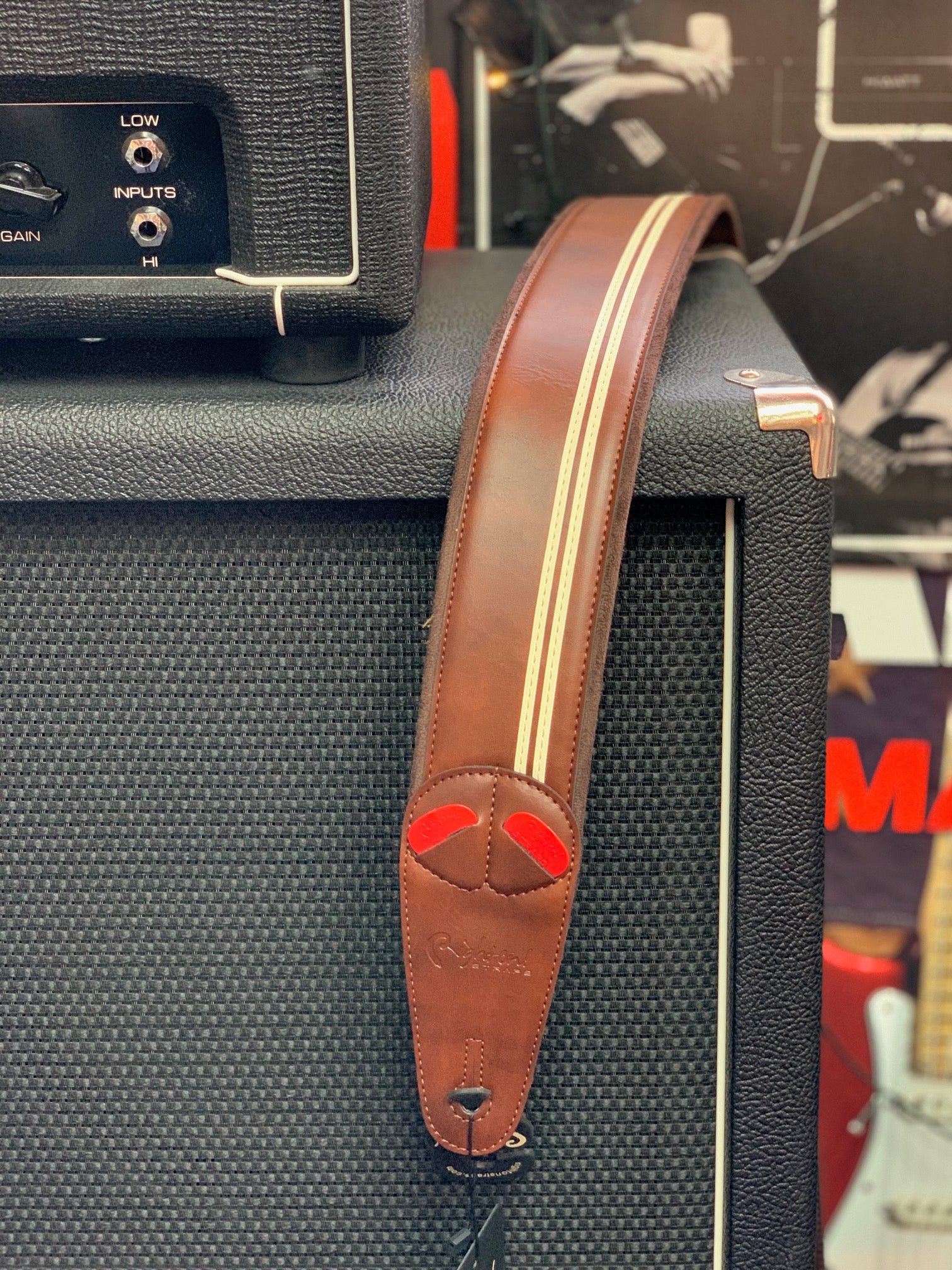 Brown leather guitar strap with tan racing stripes draped over a black amplifier cabinet, with visible control inputs and a guitar in the background.