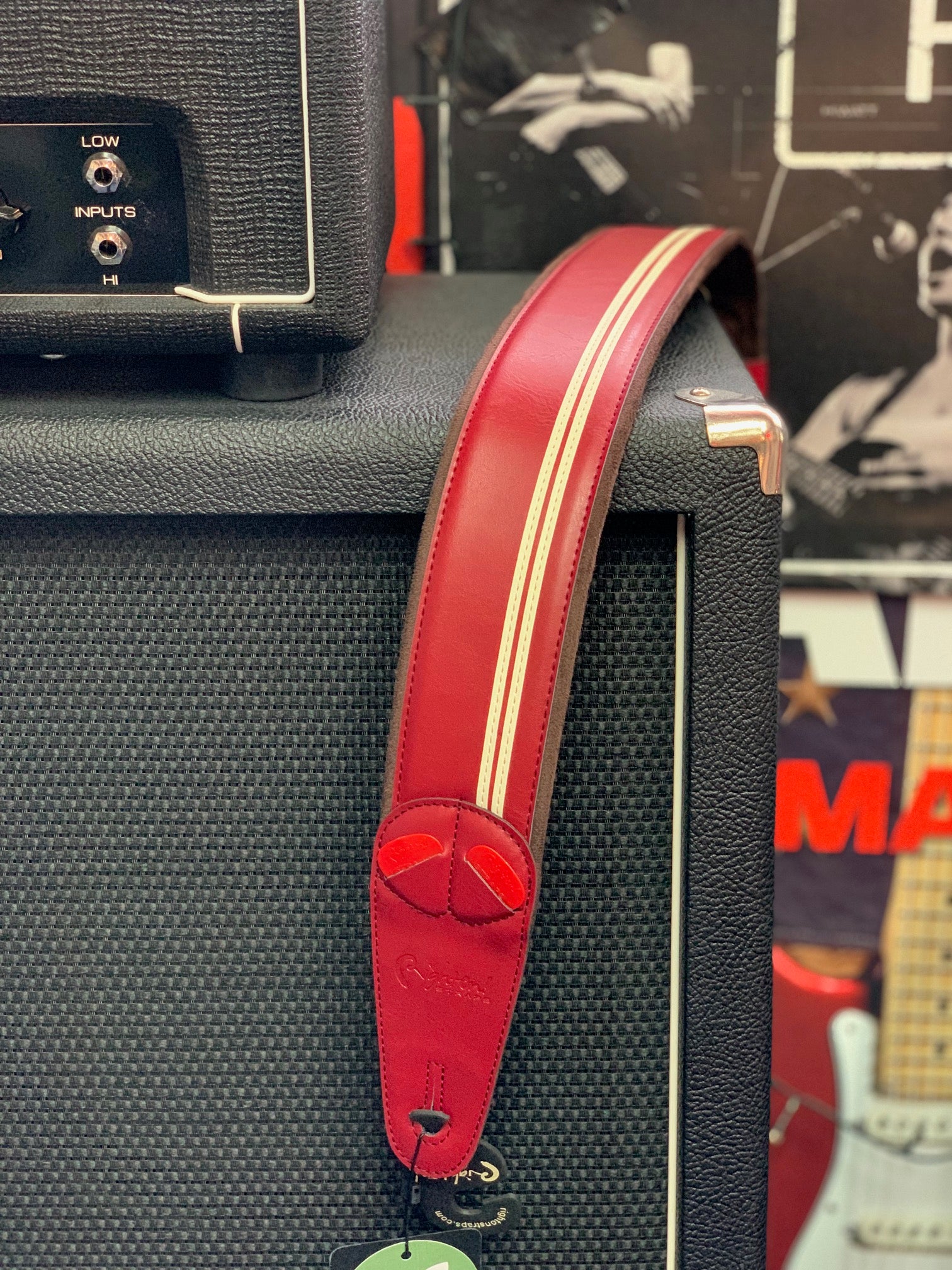 Red leather guitar strap with two white racing stripes draped over a black amplifier, electric guitar and music posters in the background.