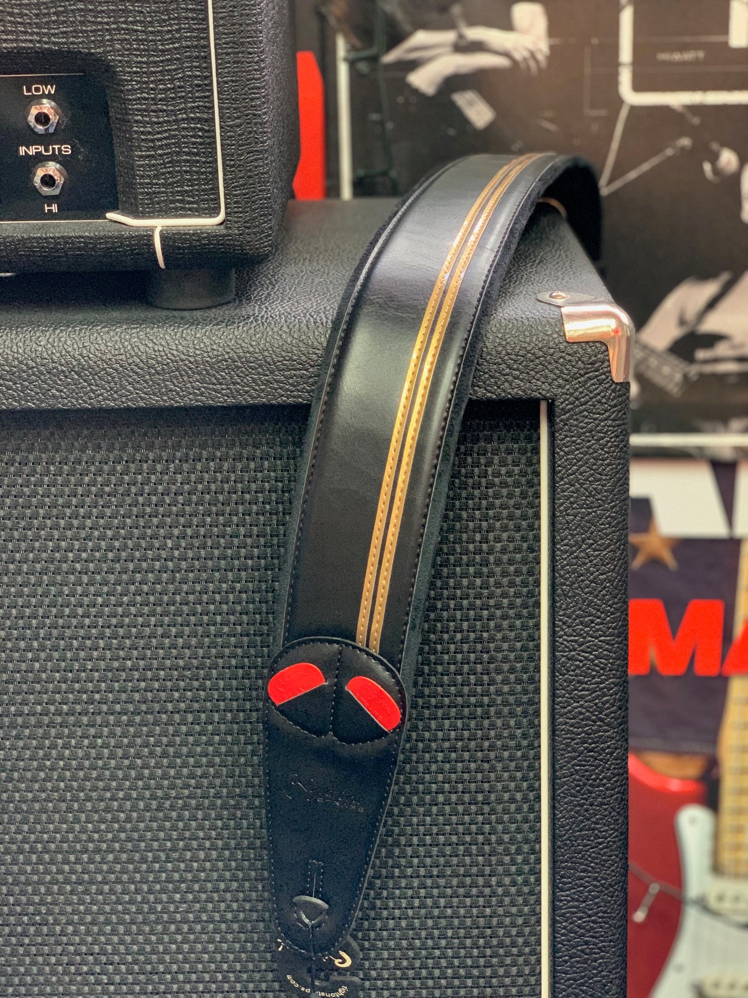 Black leather guitar strap with tan stripes and red accents rests on a textured black amplifier cabinet with visible control inputs.