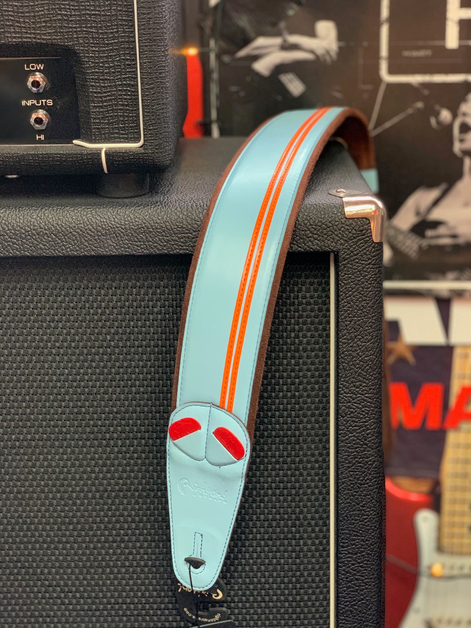 A light blue leather guitar strap with orange stripes and red accents hangs over a black amplifier.