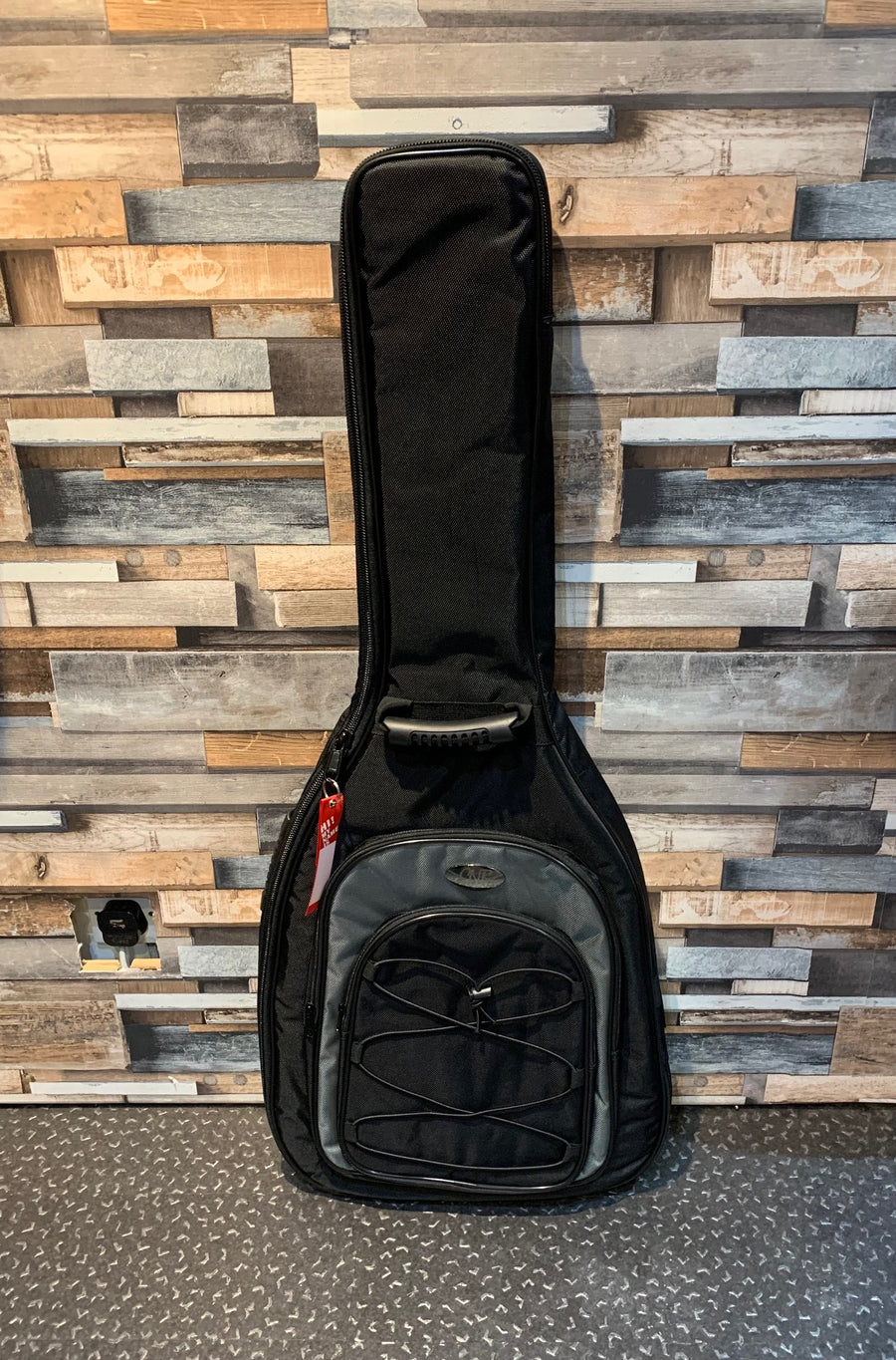 Black soft guitar case with front zip pocket, carrying handle, and adjustable bungee cord, standing upright against a wooden-patterned wall.