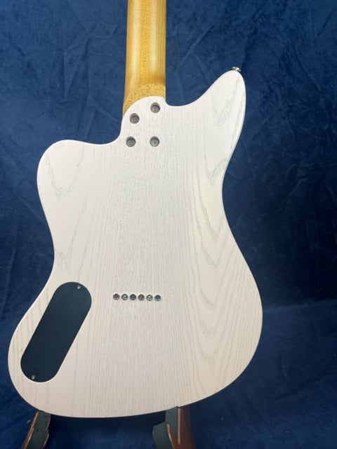 White electric guitar shown from the back, wood grain visible, with six string holes and a black electronics cover near the lower edge.
