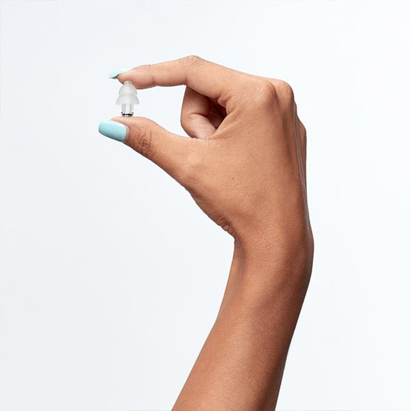 A hand with light blue nail polish holds a small, transparent earplug accessory between the thumb and index finger against a plain background.