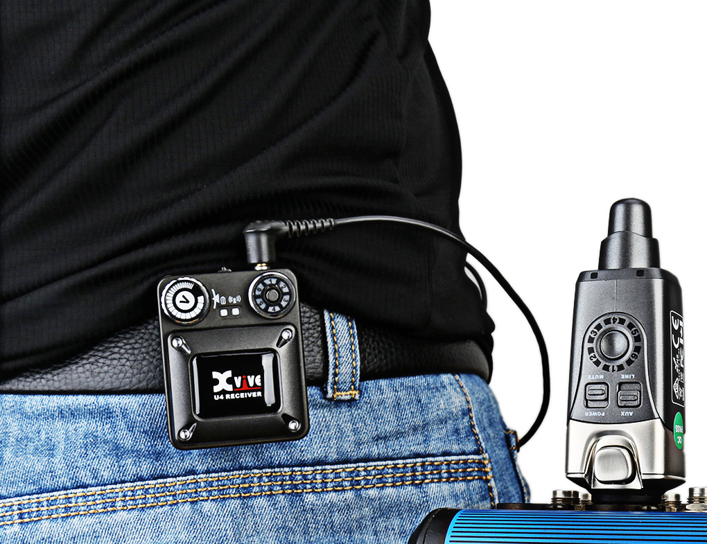 A wireless audio transmitter is clipped to a person's belt, connected by cable to a receiver attached to a nearby electronic device.