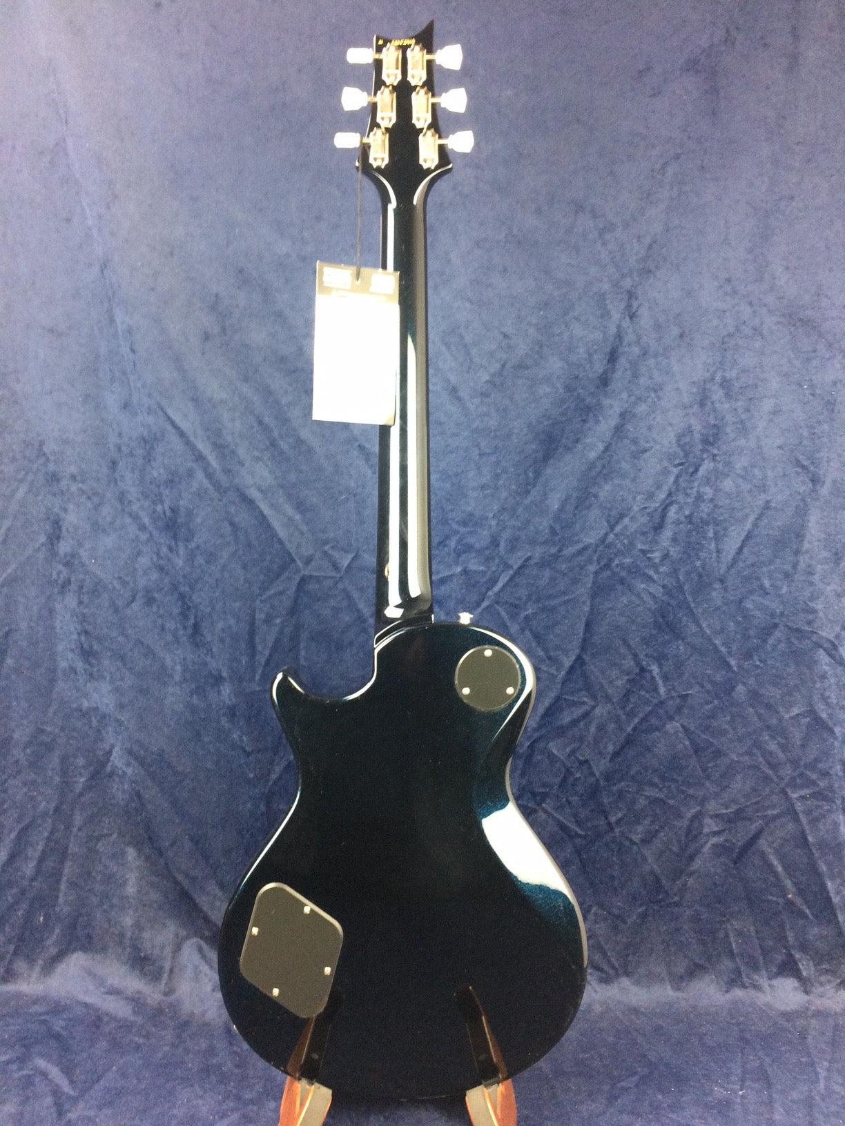Black electric guitar shown from the back with chrome tuners, round and square cover plates, and a tag hanging from the headstock against a blue backdrop.