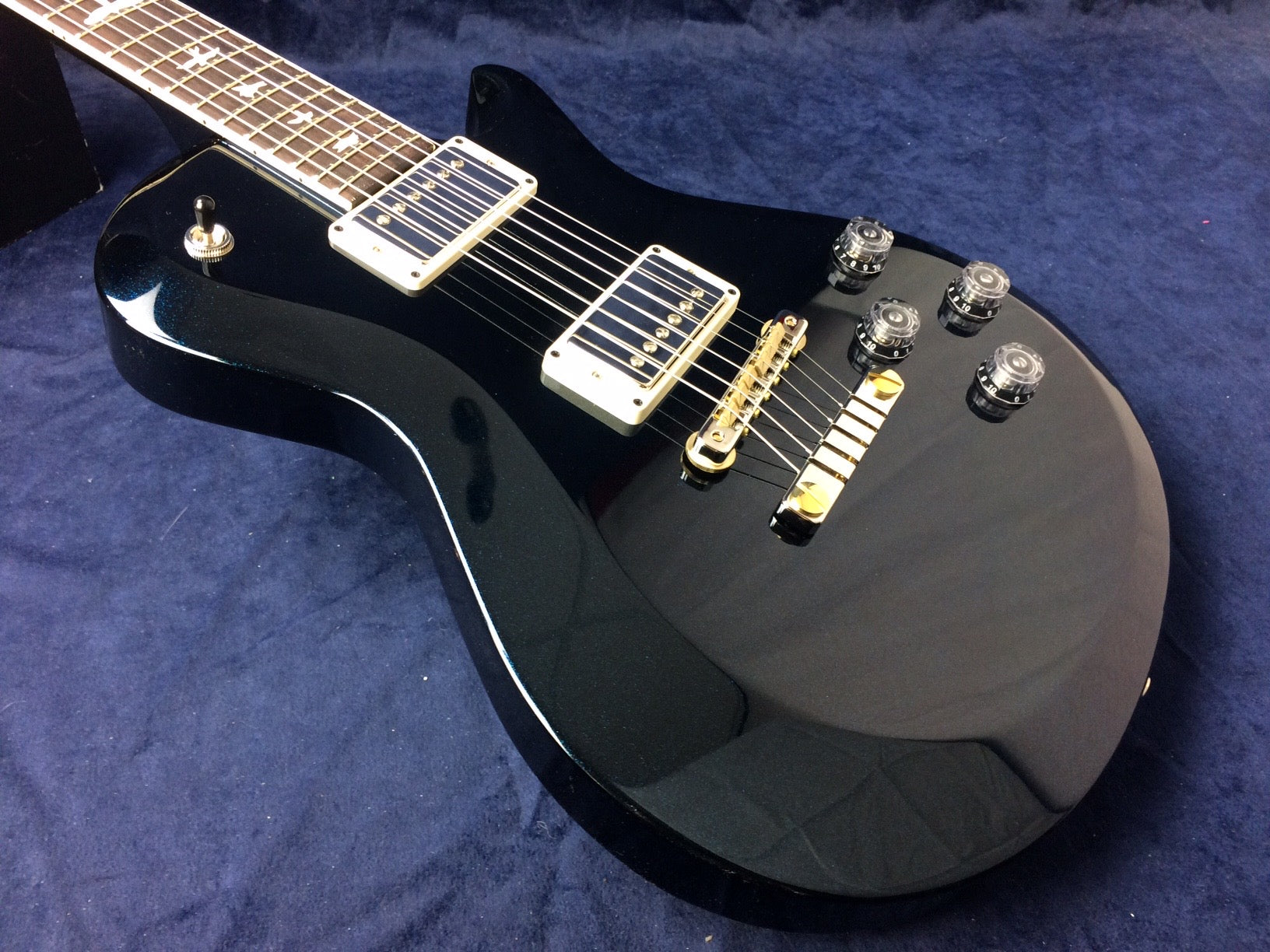Glossy black electric guitar with gold bridge, two chrome pickups, four clear control knobs, and toggle switch on a dark blue textured background.