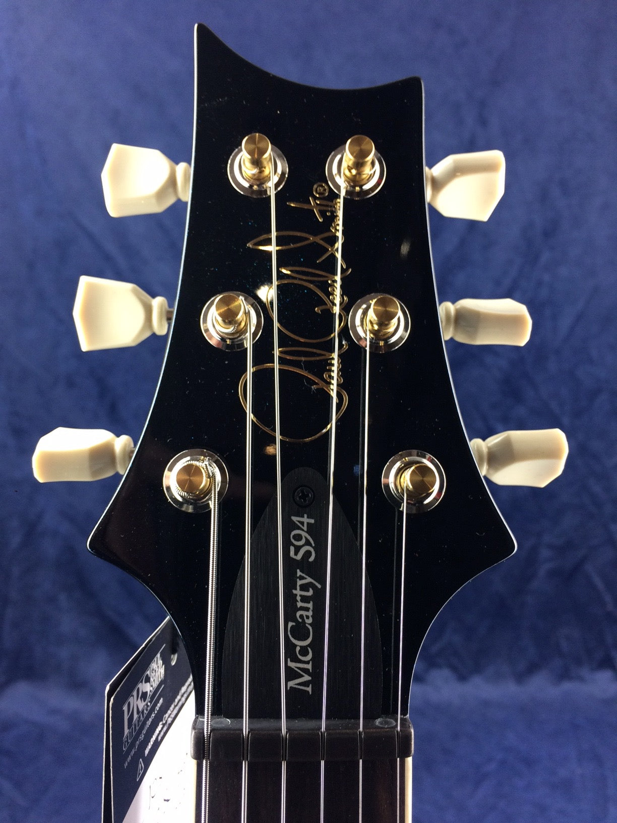Black electric guitar headstock with gold tuning pegs, white knobs, and "McCarty 594" plus a gold signature on the front.