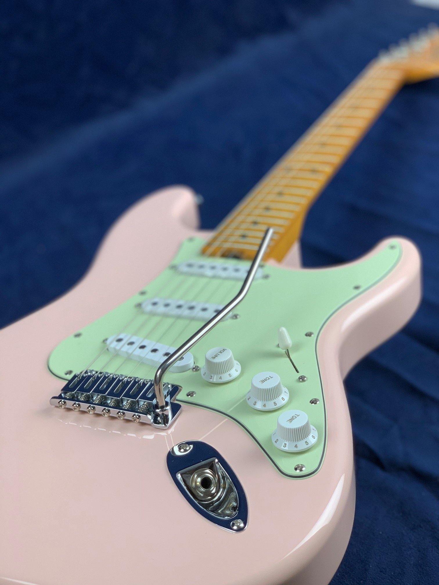 Electric guitar with a pastel pink body, mint green pickguard, white knobs, and a maple neck, shown lying on a dark blue fabric background.