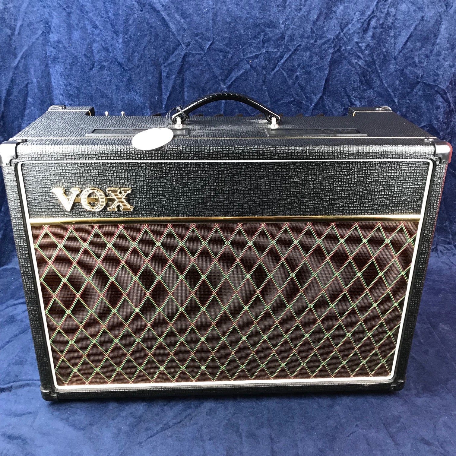 Amplificateur VOX noir avec poignée sur le dessus et grille avant marron ornée de losanges verts et rouges.