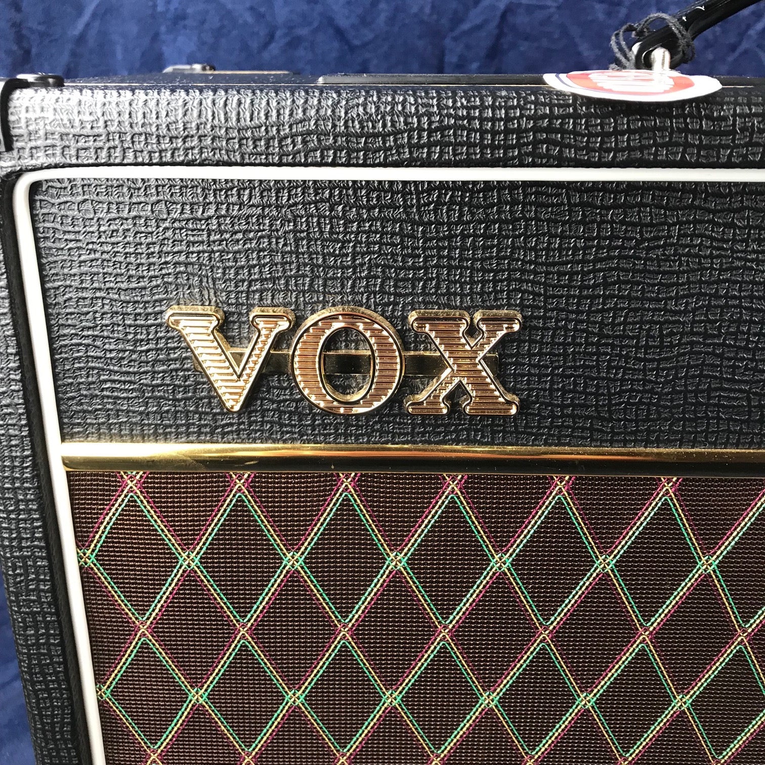 Logo doré VOX sur un ampli à revêtement noir texturé et grille marron à motifs losanges verts et rouges.