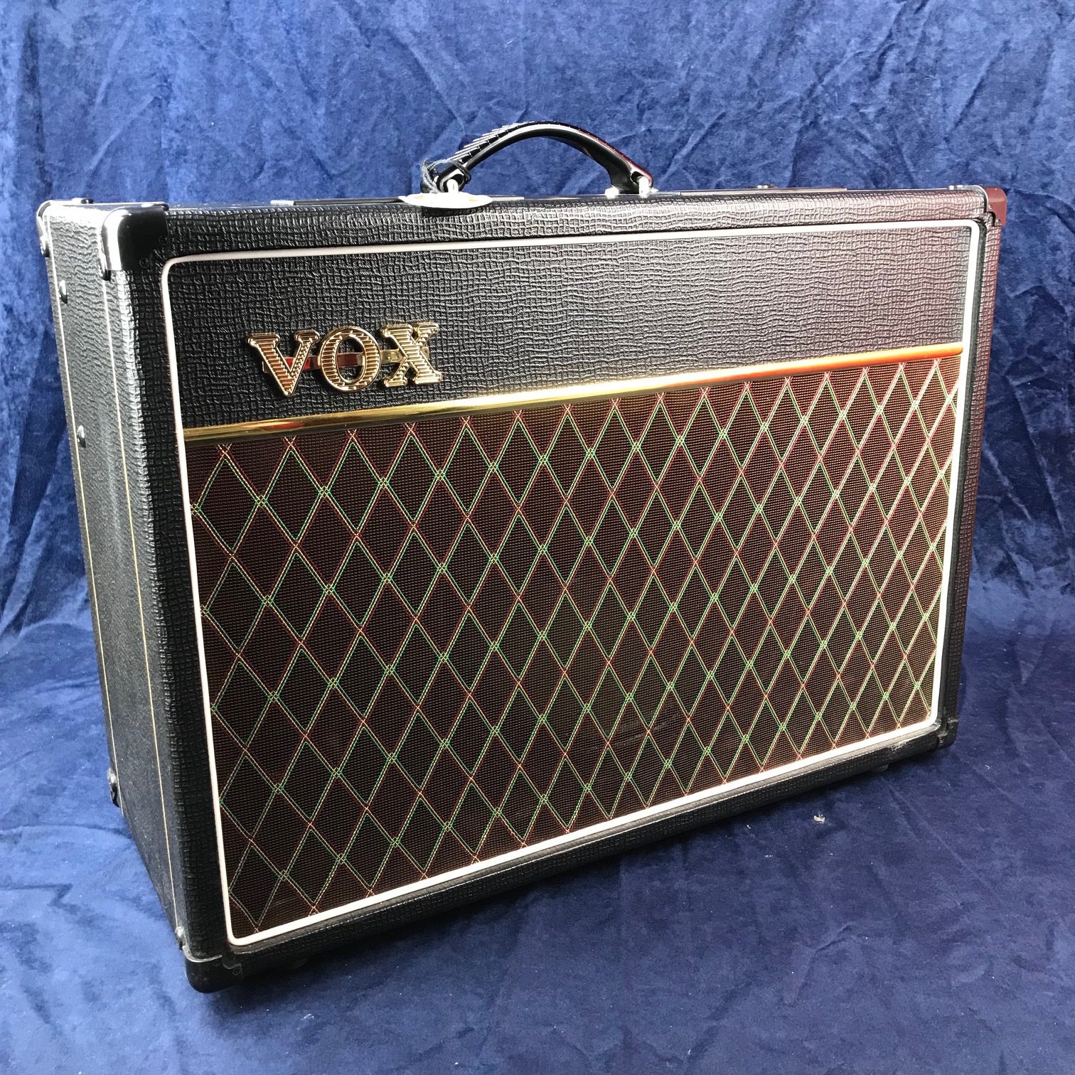 Amplificateur Vox rectangulaire, grille avant marron avec motif losange multicolore, logo doré en haut à gauche, poignée noire sur le dessus.