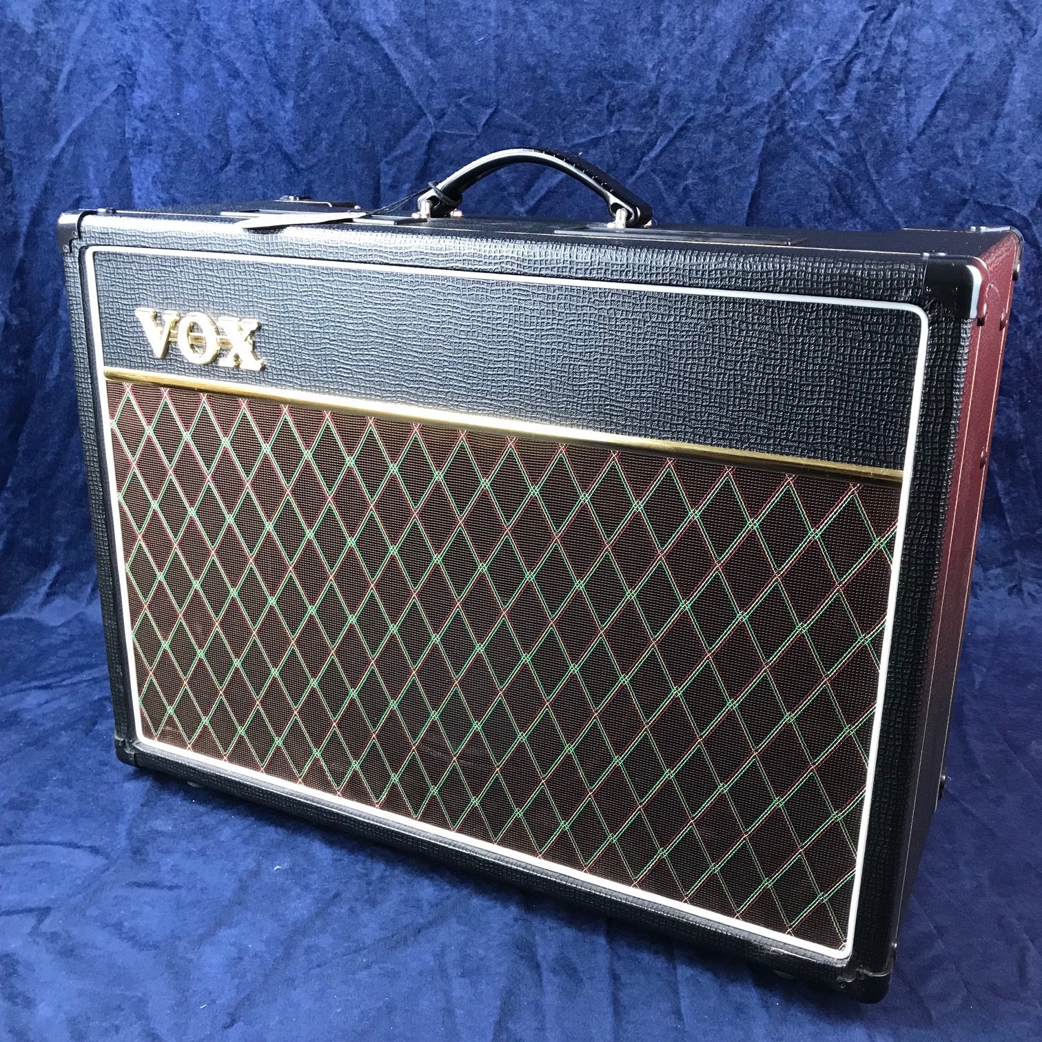 Amplificateur Vox noir avec grille marron ornée d’un motif losange vert et rouge, poignée sur le dessus, logo doré en haut à gauche.