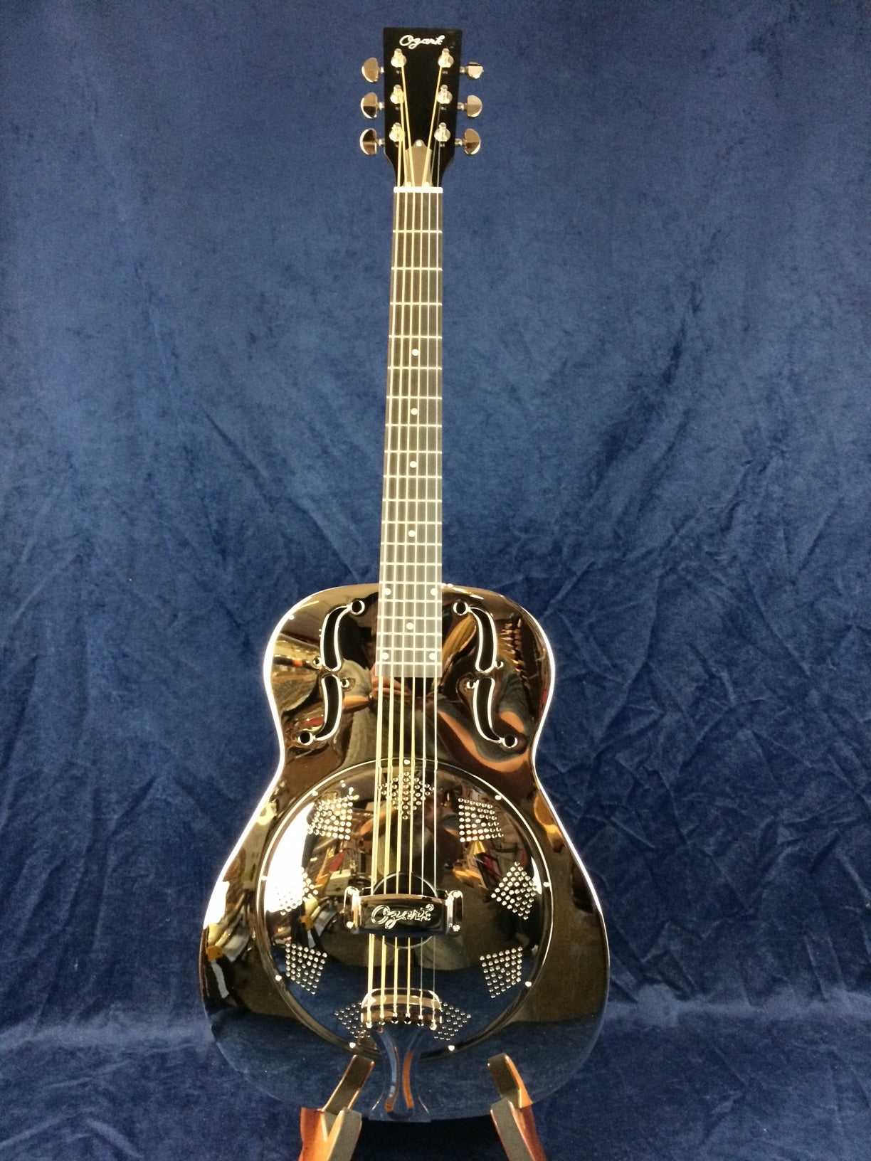 Shiny metal body resonator guitar with intricate grill pattern and two f-shaped sound holes, six strings, and the Ozark logo on the headstock.