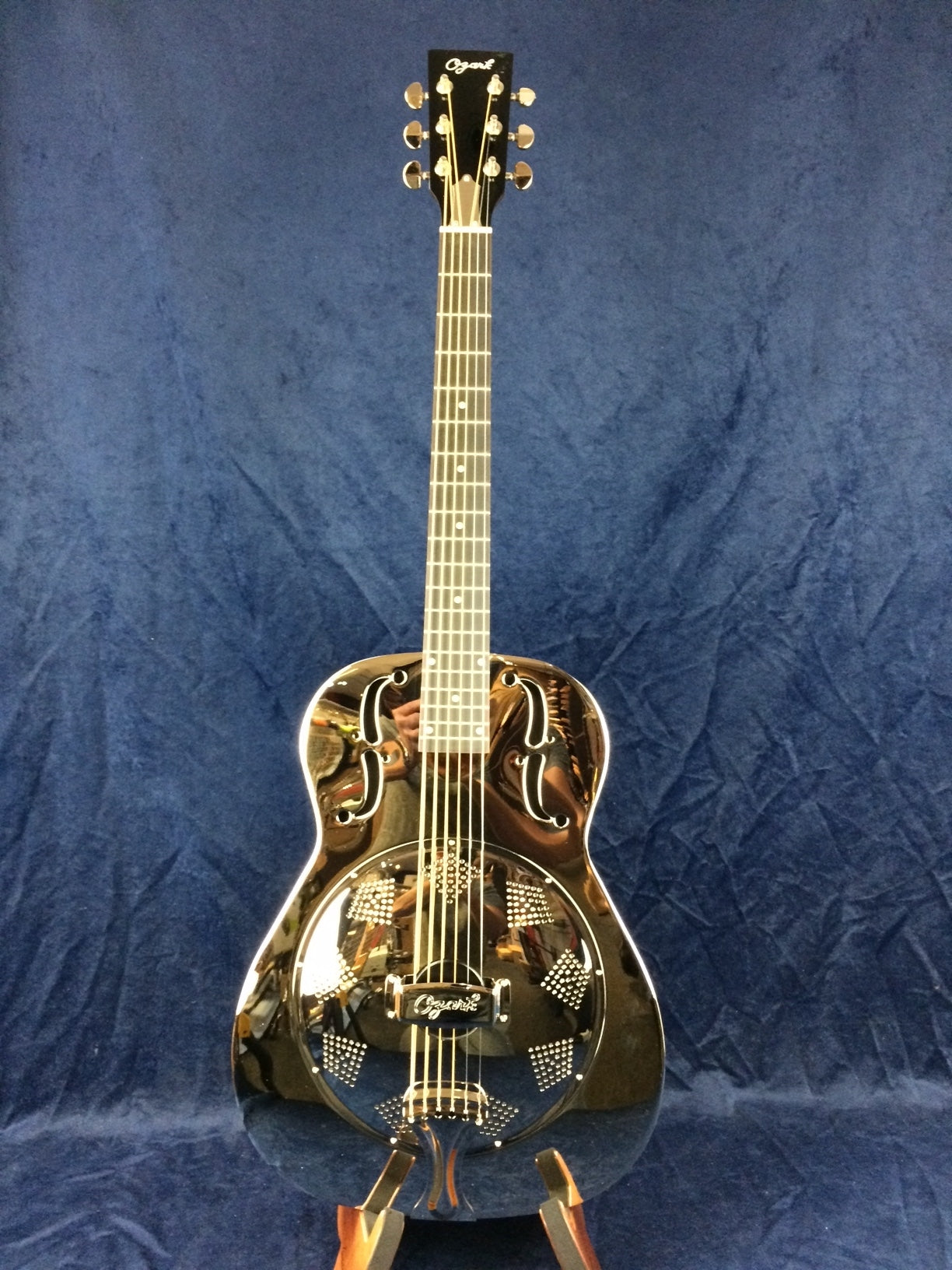 Shiny metal-bodied resonator guitar with decorative sound holes and a circular, perforated cover plate on the front, standing upright on a stand.