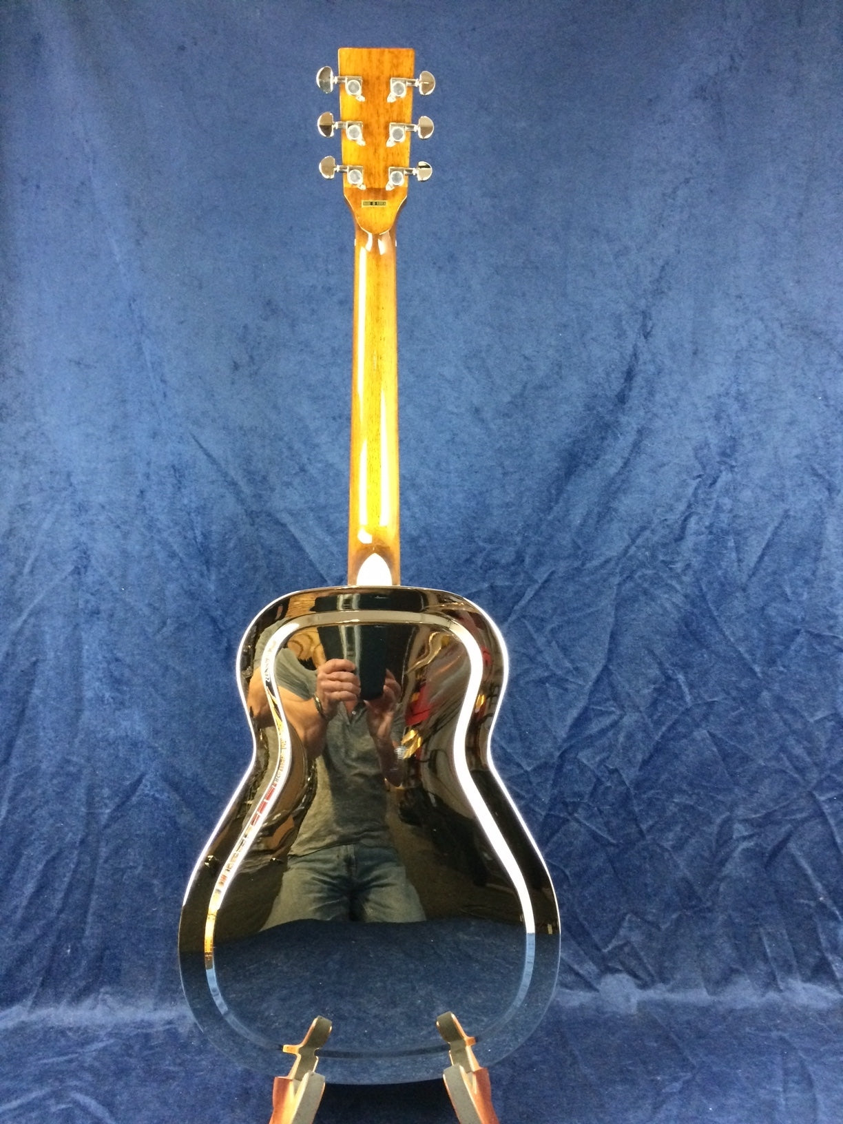 A resonator guitar with a shiny, reflective metal back and a wooden neck, standing on a blue textured background. A person’s reflection is visible.