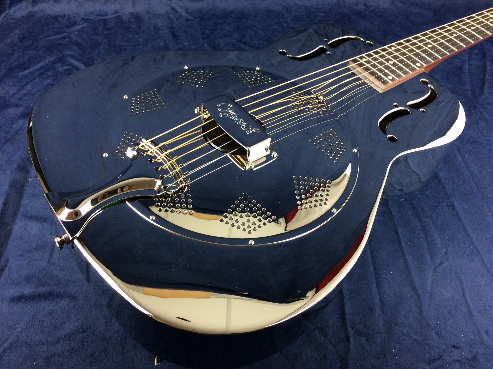 Shiny metal resonator guitar with decorative perforated patterns, f-shaped sound holes, six strings, and a reflective dark blue body.