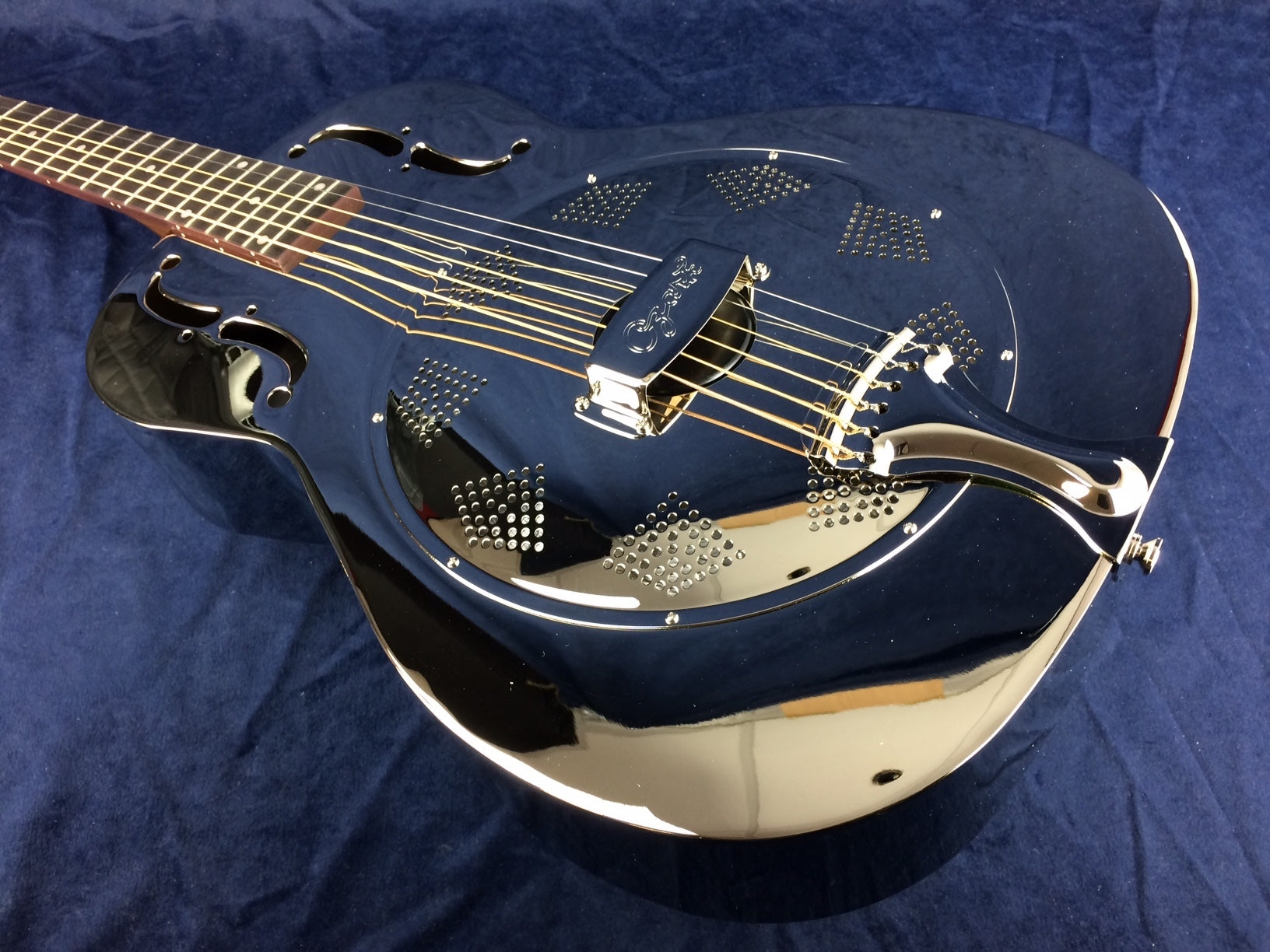 Shiny metal resonator guitar with six strings, decorative sound holes, and circular perforated patterns on the body, resting on a dark blue surface.