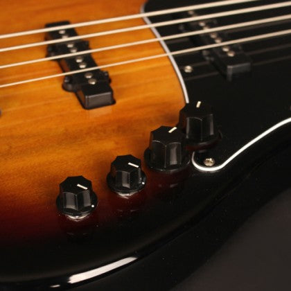 Close-up of an electric bass guitar showing four control knobs on a glossy black and sunburst body with metal strings and pickups visible.