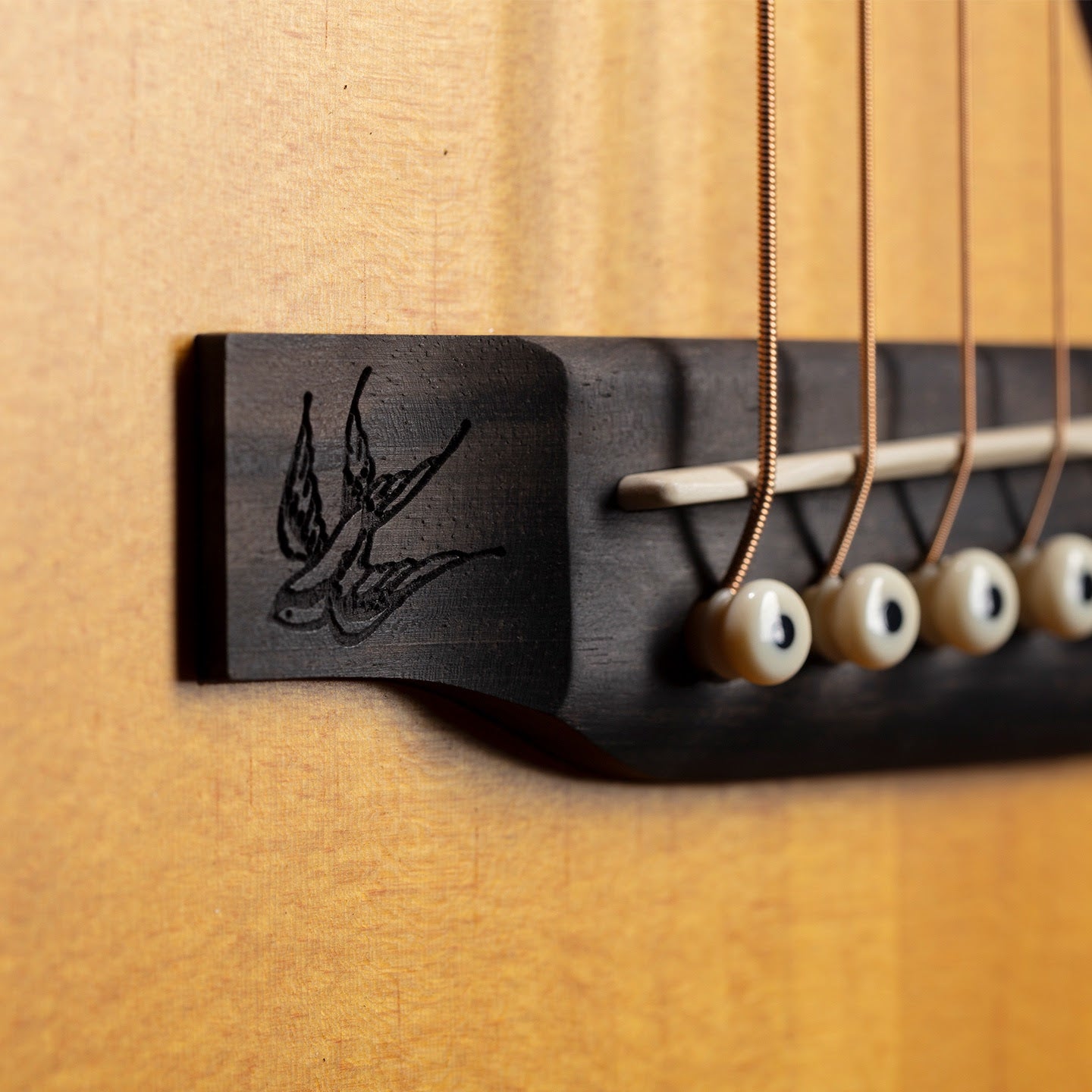 Close-up of an acoustic guitar bridge with a swallow bird design engraved on the dark wood, showing five metal strings and white bridge pins.