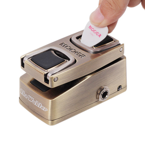 A hand inserts a guitar pick into a compact metallic effects pedal labeled "The Wahter" by Mooer, with black rubber pads and a side input jack.